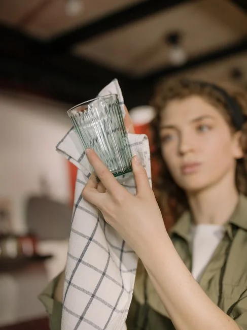 expertise-01 Jeune femme concentrée aux cheveux bouclés, agenouillée au sol et rangeant des vêtements dans des cartons avant d’emménager dans un nouvel appartement